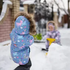 Dzieci w zimowych kurtkach bawią się na śniegu. Jedno w niebieskiej kurtce z jednorożcami, drugie w tle.