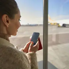Kobieta trzyma telefon i paszport, gotowa do podróży. Na ekranie telefonu widoczna jest karta pokładowa.