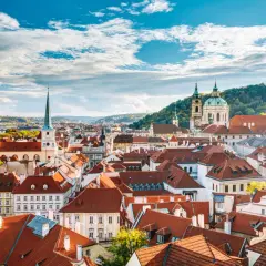 Widok na dachy Malé Strany w Pradze z wieżami kościołów i wzgórzem w tle.