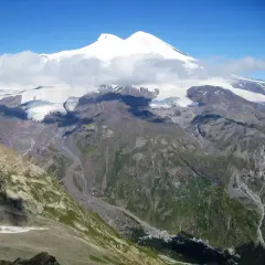 Elbrus, najwyższy szczyt Kaukazu, w chmurach, z lodowcami i zielonymi dolinami poniżej.