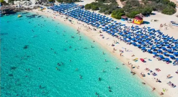 Słoneczny dzień na plaży Makronissos. Turkusowa woda, biały piasek, rzędy niebieskich parasoli i mnóstwo plażowiczów cieszących się słońcem i kąpielą.