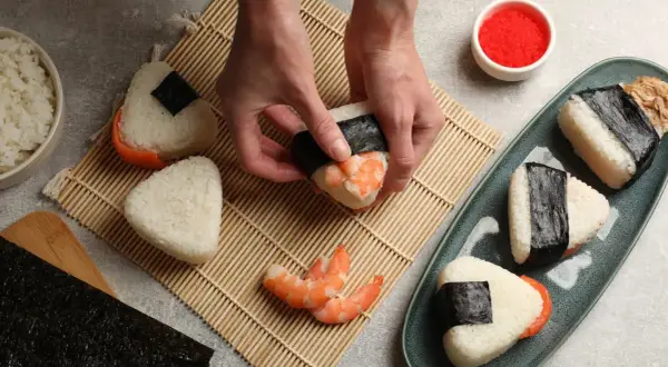 Ręce formują onigiri z krewetkami, tworząc pyszne japońskie jedzenie. Obok miska ryżu i talerz z gotowymi przysmakami.