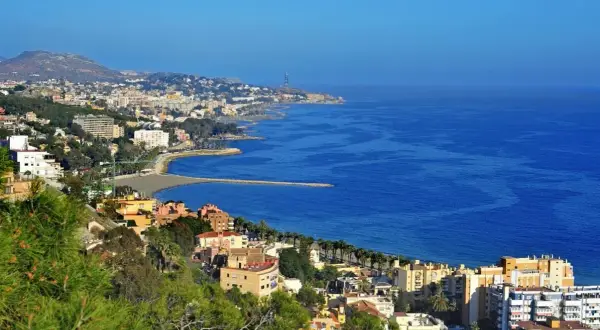Widok na malaga plaże i miasto z zielonymi wzgórzami w tle. Błękitne morze lśni w słońcu.
