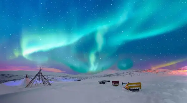 Niesamowita zorza polarna nad śnieżnym krajobrazem Norwegii. W oddali widać tipi i sanie, gotowe na nocną przygodę.