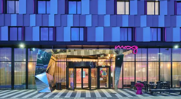 Nowoczesny budynek Moxy Hotels Pyrzowice nocą, z niebieską fasadą i neonowym logo.
