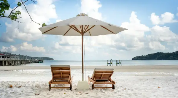 Dwa leżaki pod parasolem na piaszczystej plaży, z widokiem na spokojne morze i błękitne niebo.