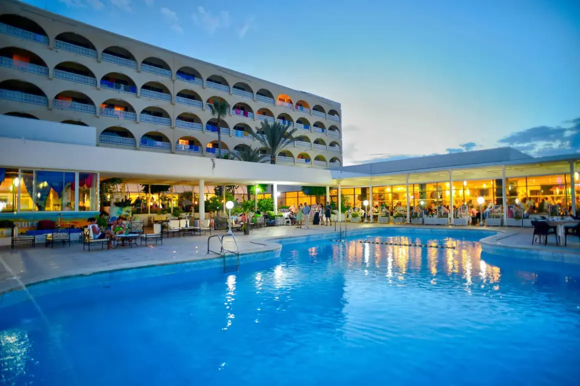 Basen i restauracja hotelowa wieczorem. Wiele osób siedzi przy stolikach. Opinie o tym resorcie są pozytywne.