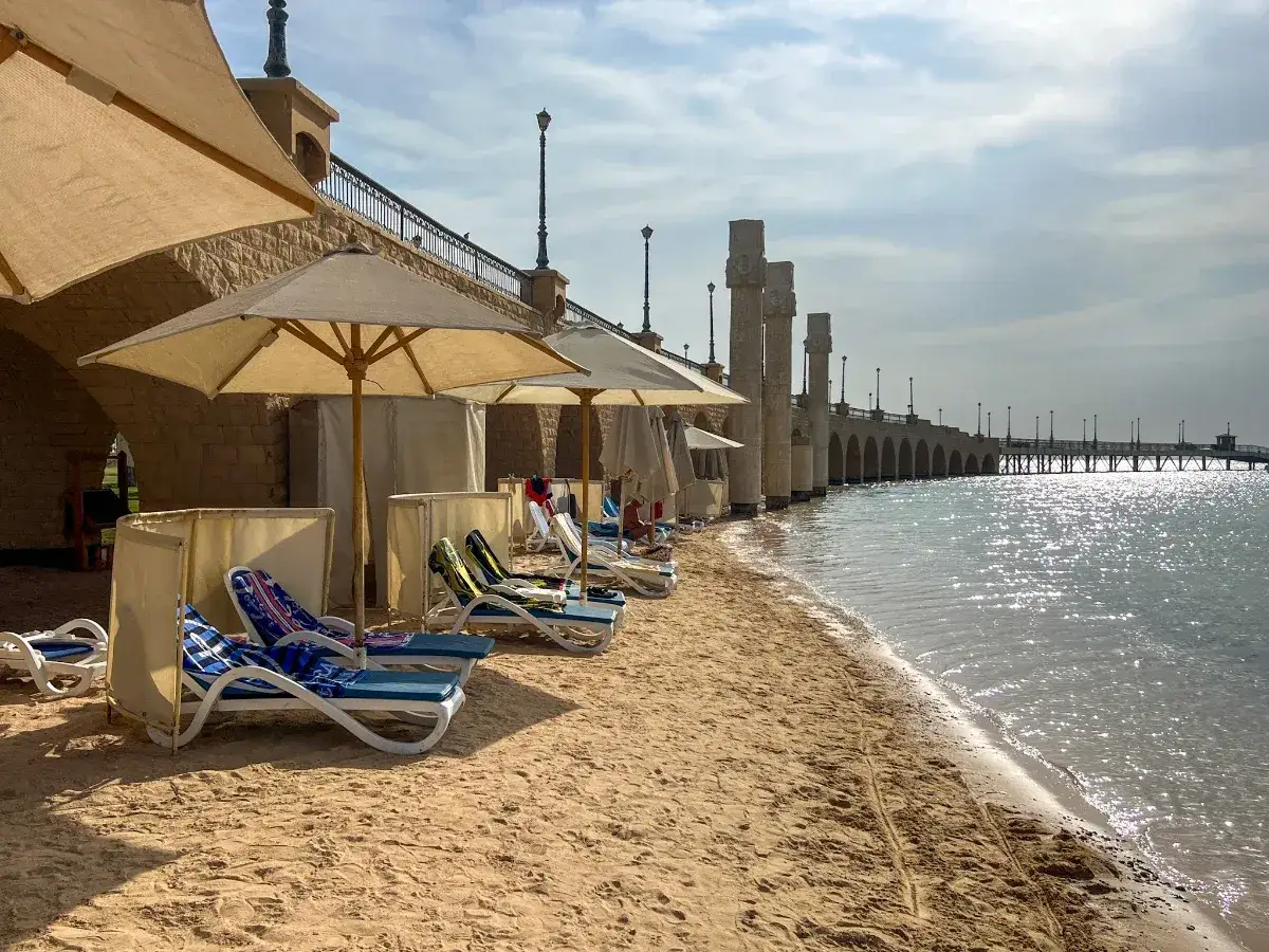 Plaża w Egipcie, leżaki pod parasolami, most nad wodą. Czy jest bezpiecznie? Tak, wygląda na spokojny wypoczynek.