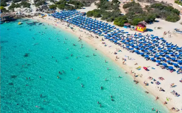 Słoneczny dzień na plaży Makronissos. Turkusowa woda, biały piasek, rzędy niebieskich parasoli i mnóstwo plażowiczów cieszących się słońcem i kąpielą.