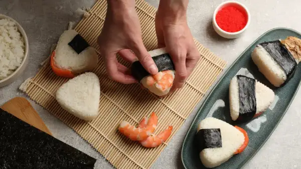 Ręce formują onigiri z krewetkami, tworząc pyszne japońskie jedzenie. Obok miska ryżu i talerz z gotowymi przysmakami.