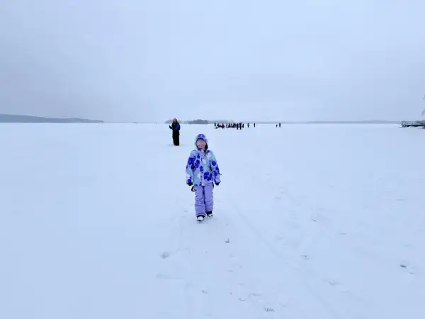 Dziewczynka w kolorowym kombinezonie na tle zamarzniętego jeziora. W oddali widać grupę ludzi. To niezapomniana laponia wycieczka.