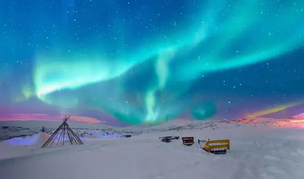 Niesamowita zorza polarna nad śnieżnym krajobrazem Norwegii. W oddali widać tipi i sanie, gotowe na nocną przygodę.