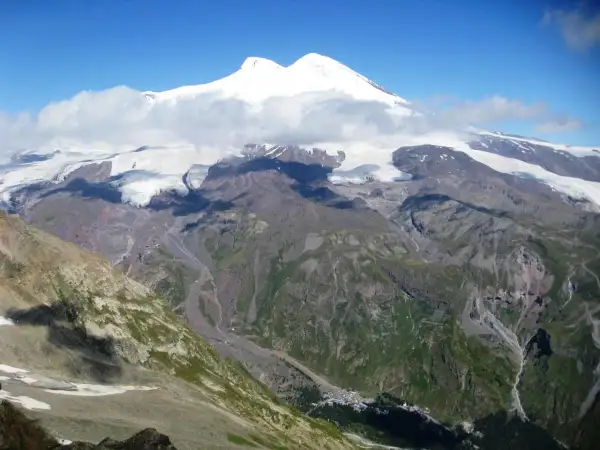 Elbrus, najwyższy szczyt Kaukazu, w chmurach, z lodowcami i zielonymi dolinami poniżej.