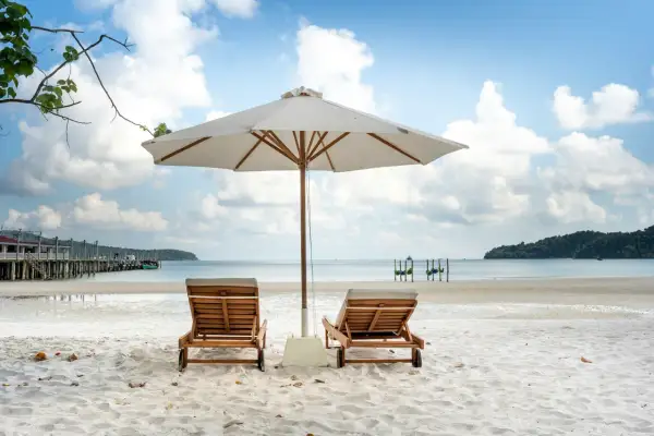 Dwa leżaki pod parasolem na piaszczystej plaży, z widokiem na spokojne morze i błękitne niebo.