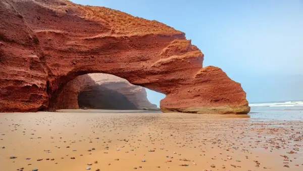 Skalny łuk na piaszczystej plaży w Maroku. Słońce wdziera się do jaskini, tworząc magiczną atmosferę.